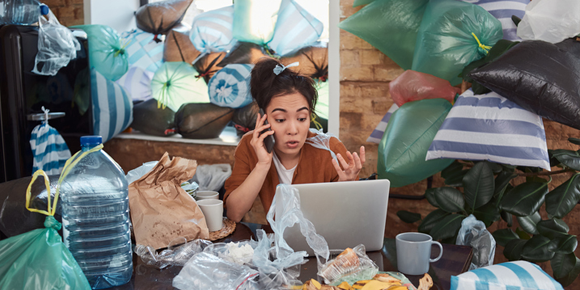 stressed woman sits at computer talking on the phone in a house filled with trash